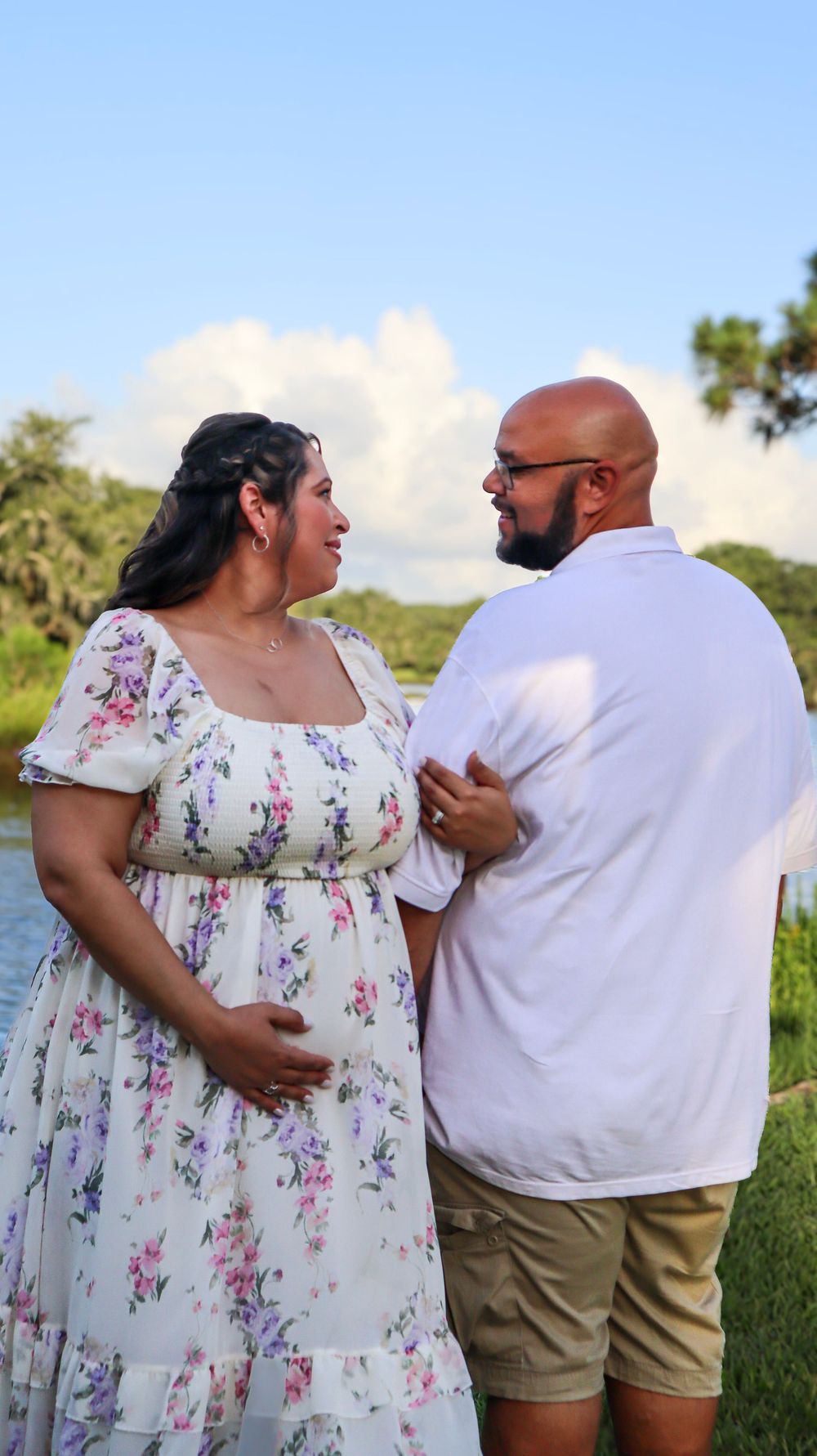 A couple shares an intimate moment by a waterfront with trees in the background.
