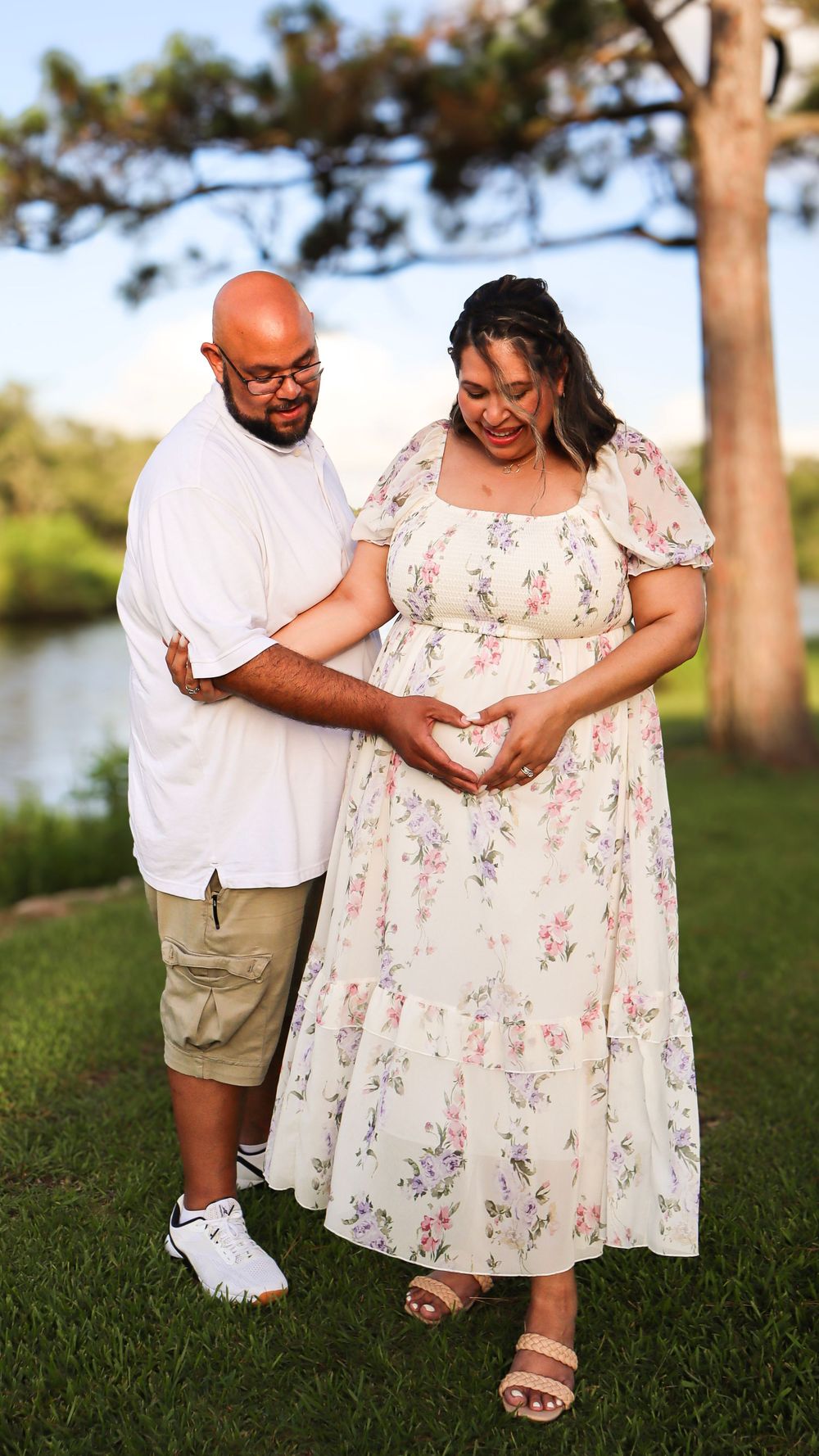 A couple forms a heart shape with their hands while posing for a maternity photo outdoors.