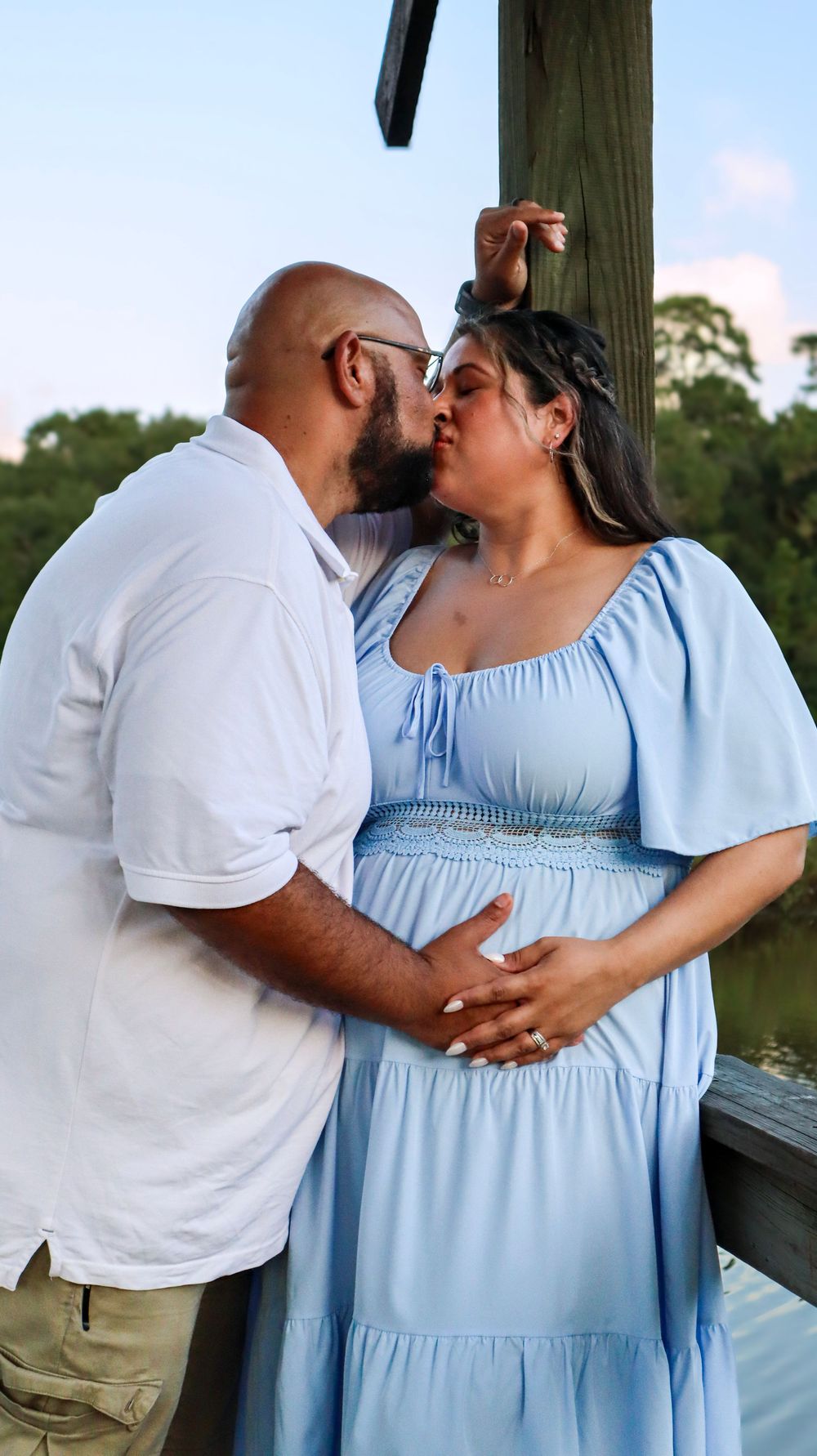 A couple shares a kiss on a wooden dock during a maternity photoshoot.