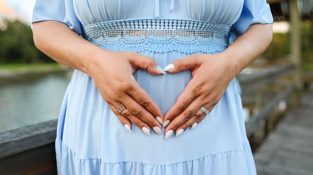 Hands form heart shape over baby bump in light blue maternity dress.