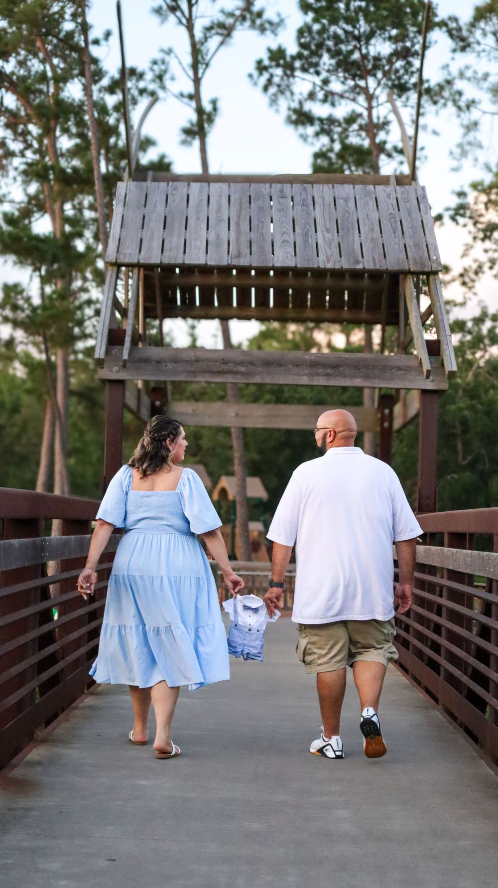 A couple walks hand in hand across a wooden bridge surrounded by trees during sunset.