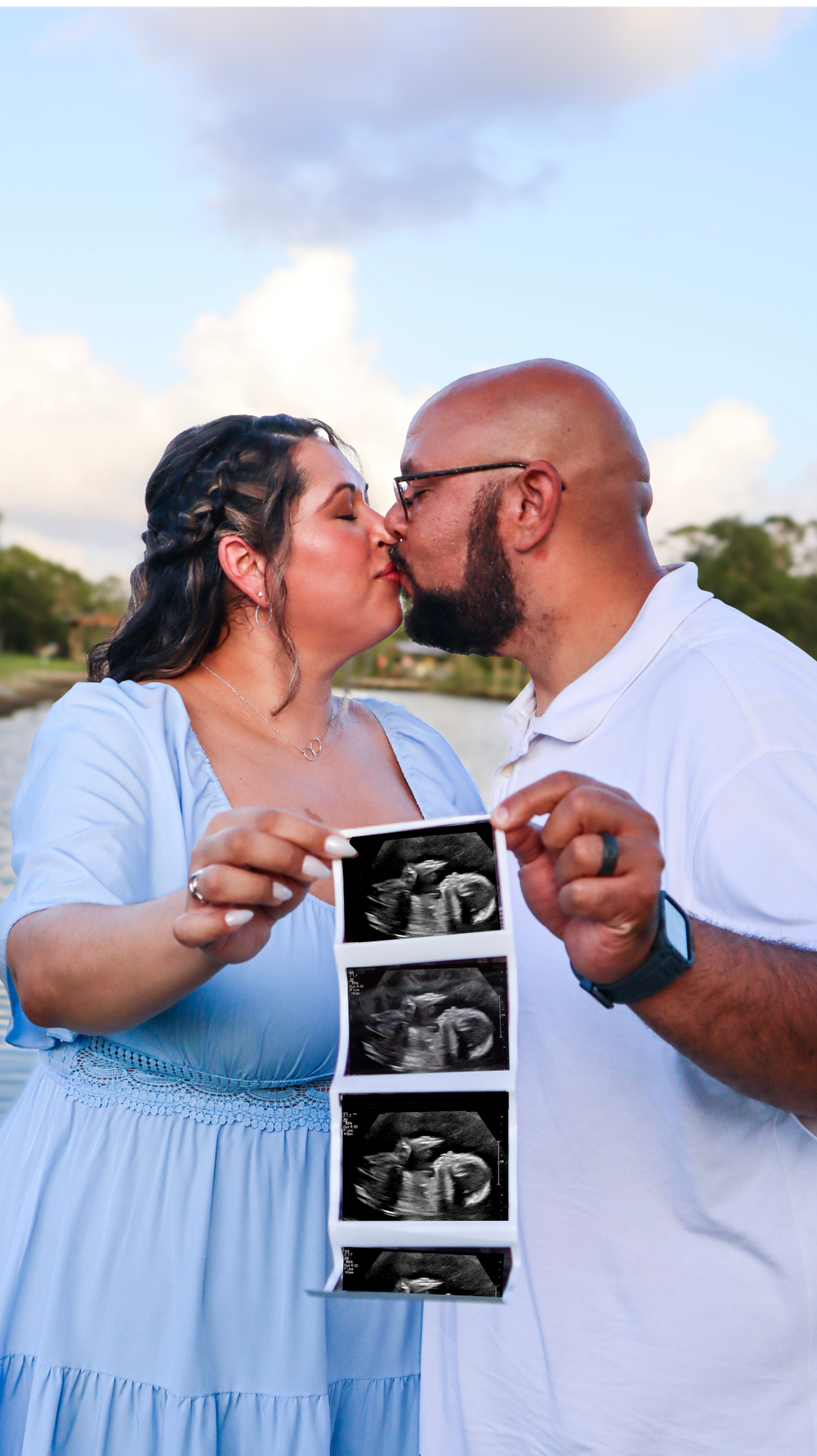 A couple in light blue and white outfits share a kiss while holding ultrasound photos during a pregnancy announcement photoshoot.