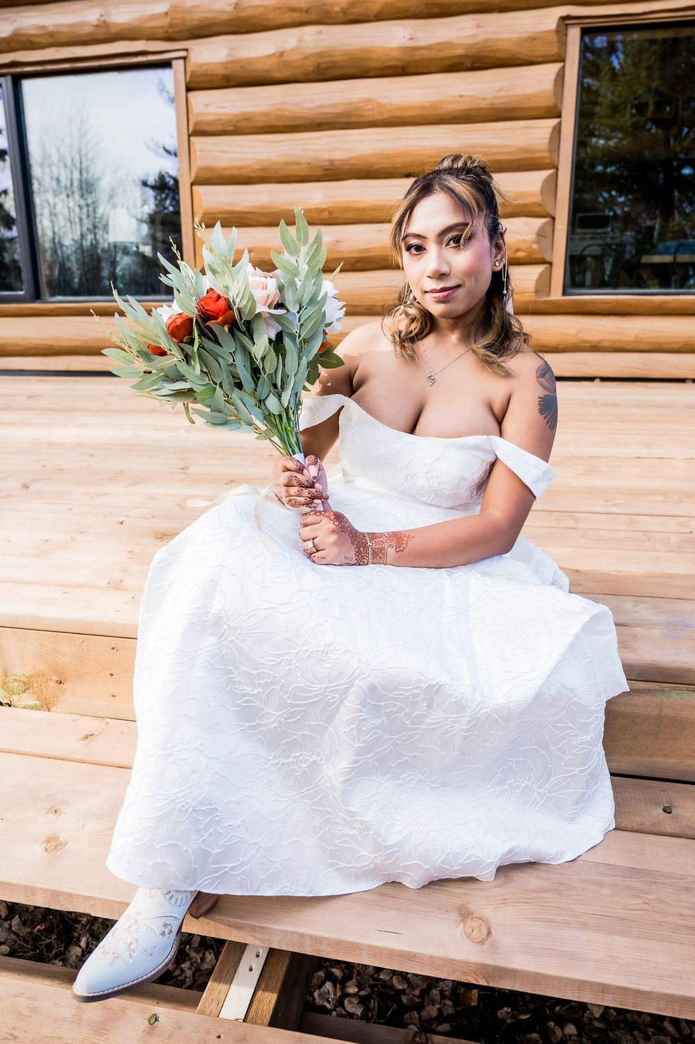 A bride in a white wedding dress sits on a rustic wooden bench outside a log cabin.