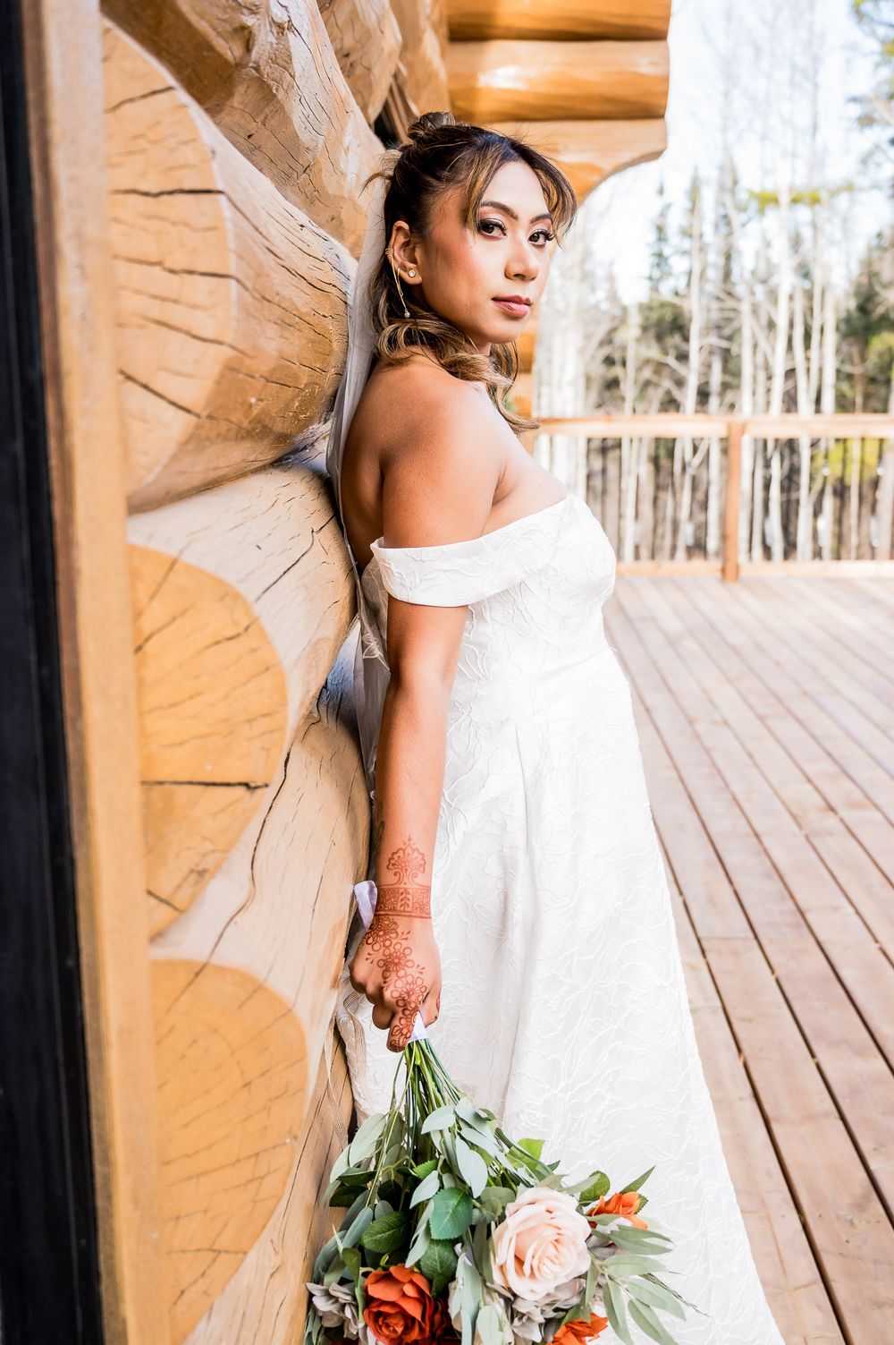 A bride in a flowing white off-shoulder gown holds a rustic bouquet while standing against a wooden log cabin wall.