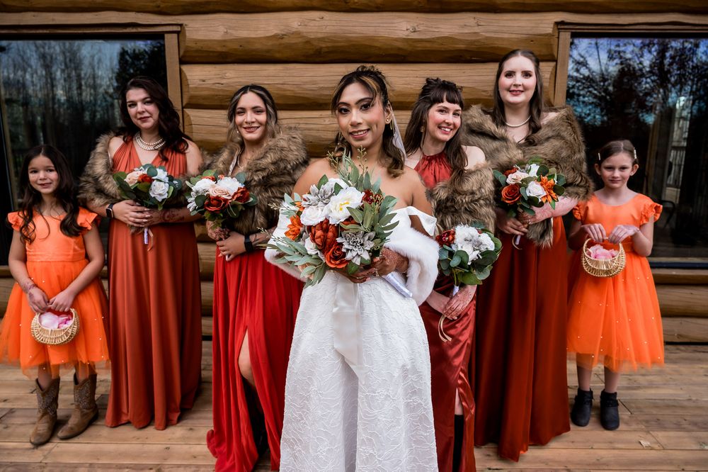 Wedding party poses outdoors wearing orange and burgundy dresses with floral bouquets at a rustic venue.