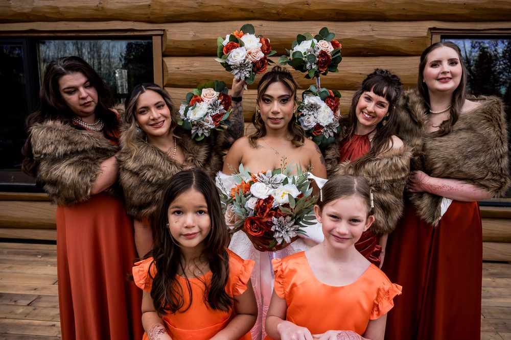 A group of bridesmaids in burnt orange dresses pose together in a rustic wooden setting.