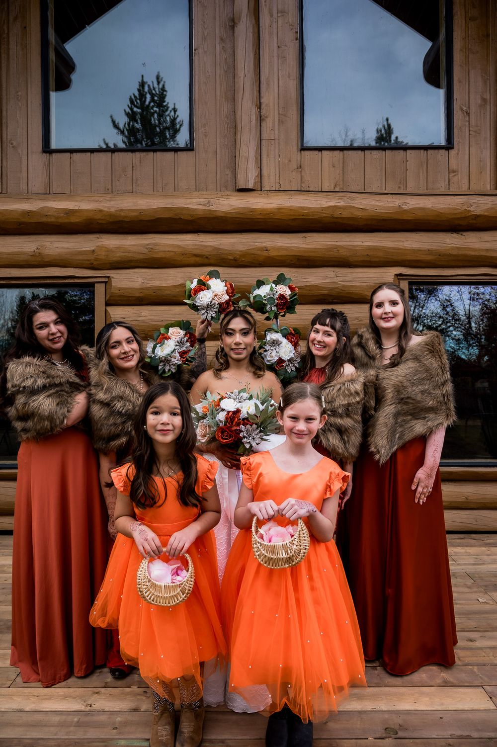 Wedding party wearing vibrant orange and burgundy dresses pose together in front of rustic wooden backdrop.