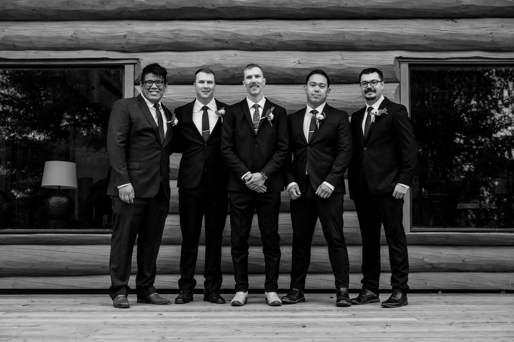 Groomsmen in black suits show off their colorful socks while sitting on a wooden porch ledge.