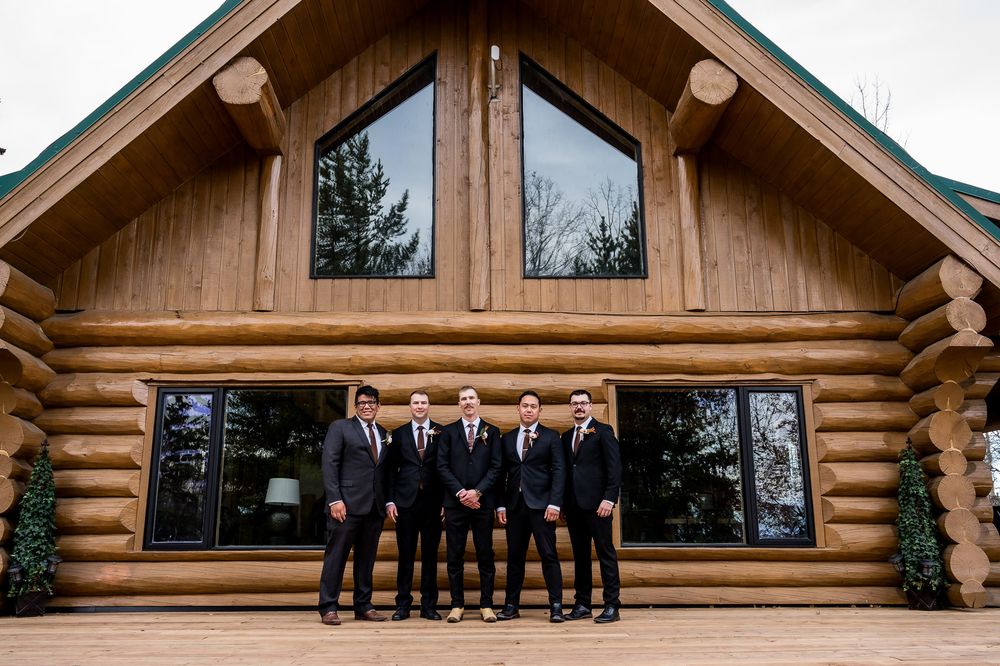 A group in formal attire poses in front of a rustic log cabin building during a wedding celebration.