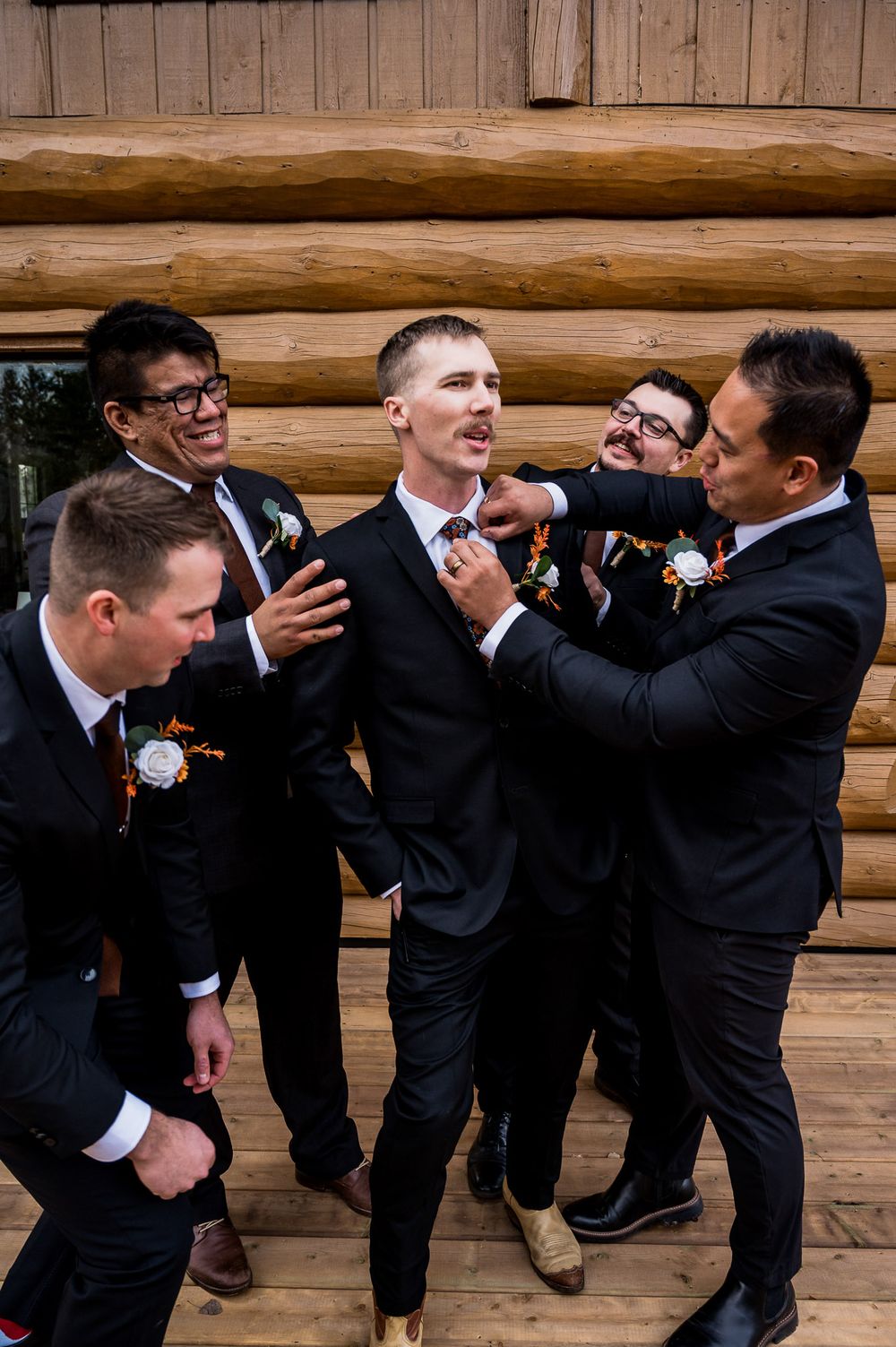 Groomsmen in black suits playfully interact and laugh together against a rustic log cabin wall background.