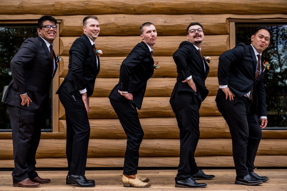 A sequence of groomsmen bending backwards in matching black suits against a wooden log cabin wall.