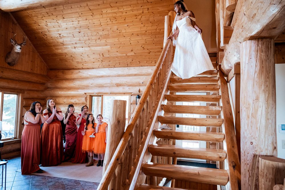 A bride in a white dress descends wooden stairs in a rustic log cabin while bridesmaids in burgundy dresses watch below.