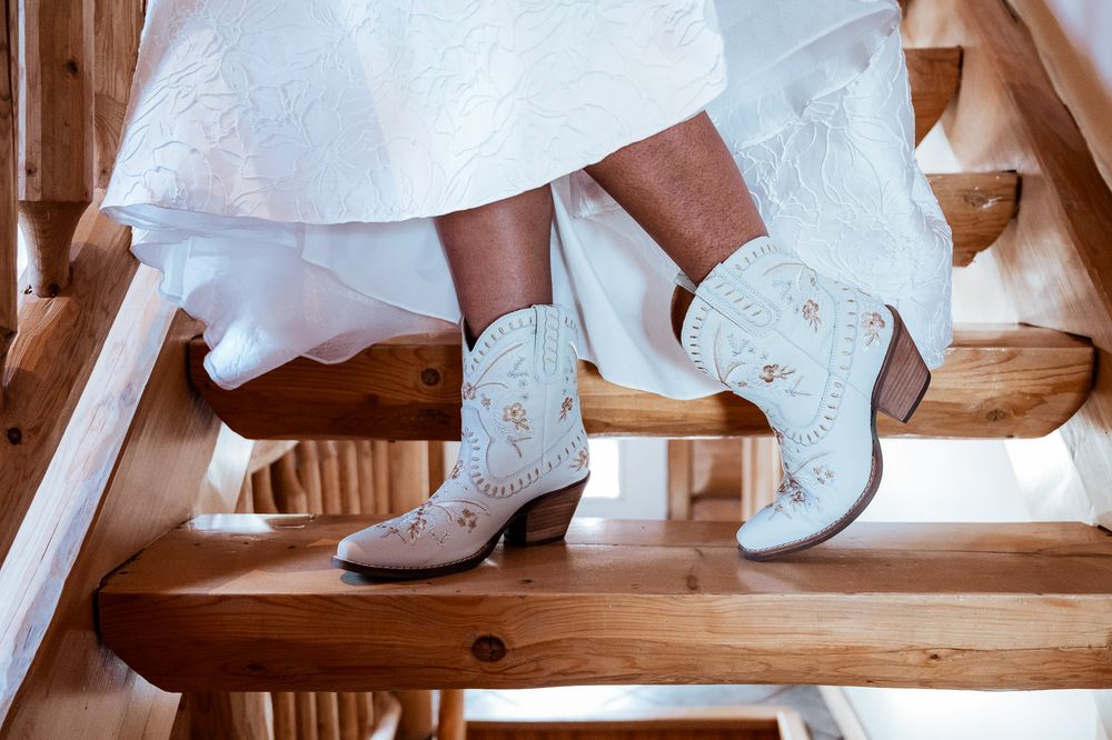 Wedding dress details showing white lace shoes and gown hem against a rustic wooden backdrop.