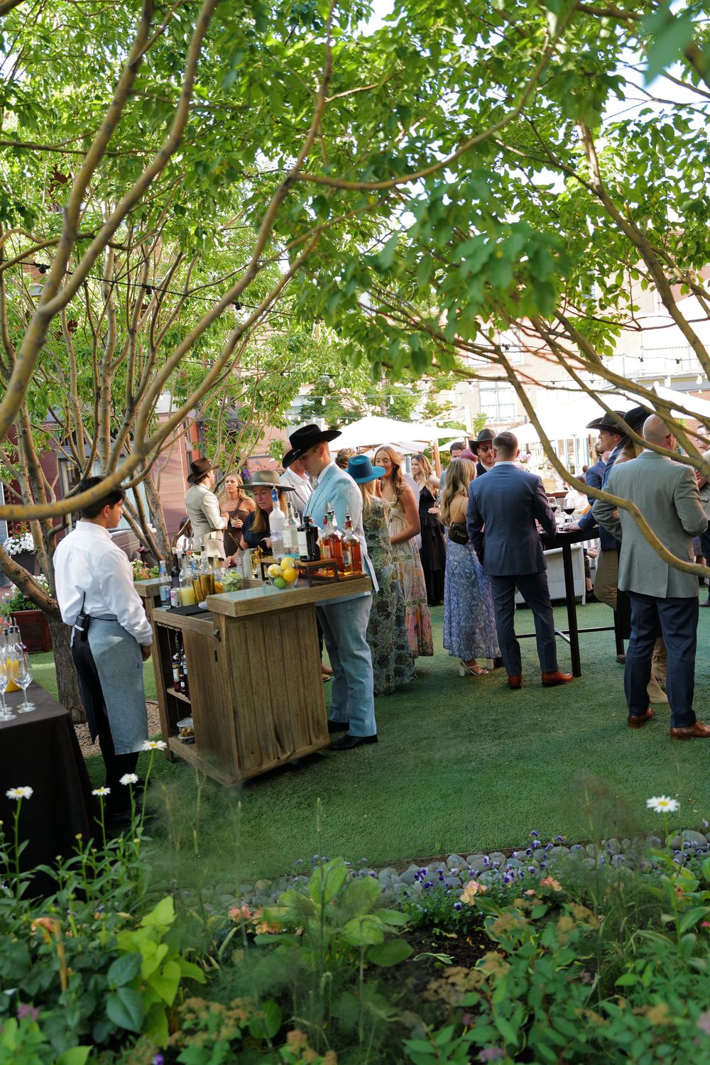People gather at an outdoor garden party under leafy trees with wooden serving stations and refreshments. aspen vibes