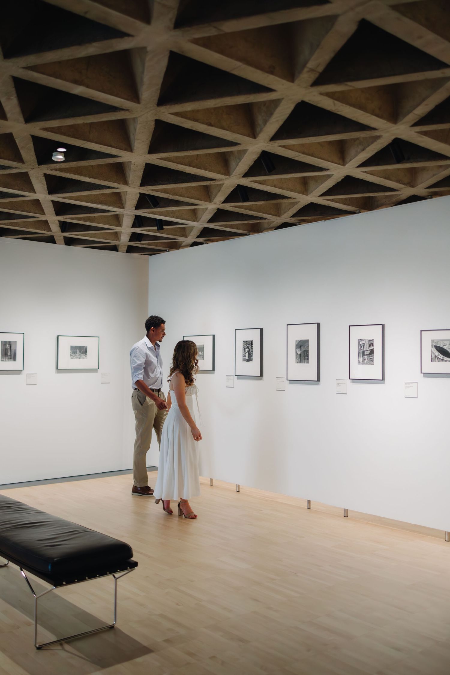Gallery visitors view black and white photographs displayed along white walls in a modern exhibition space.