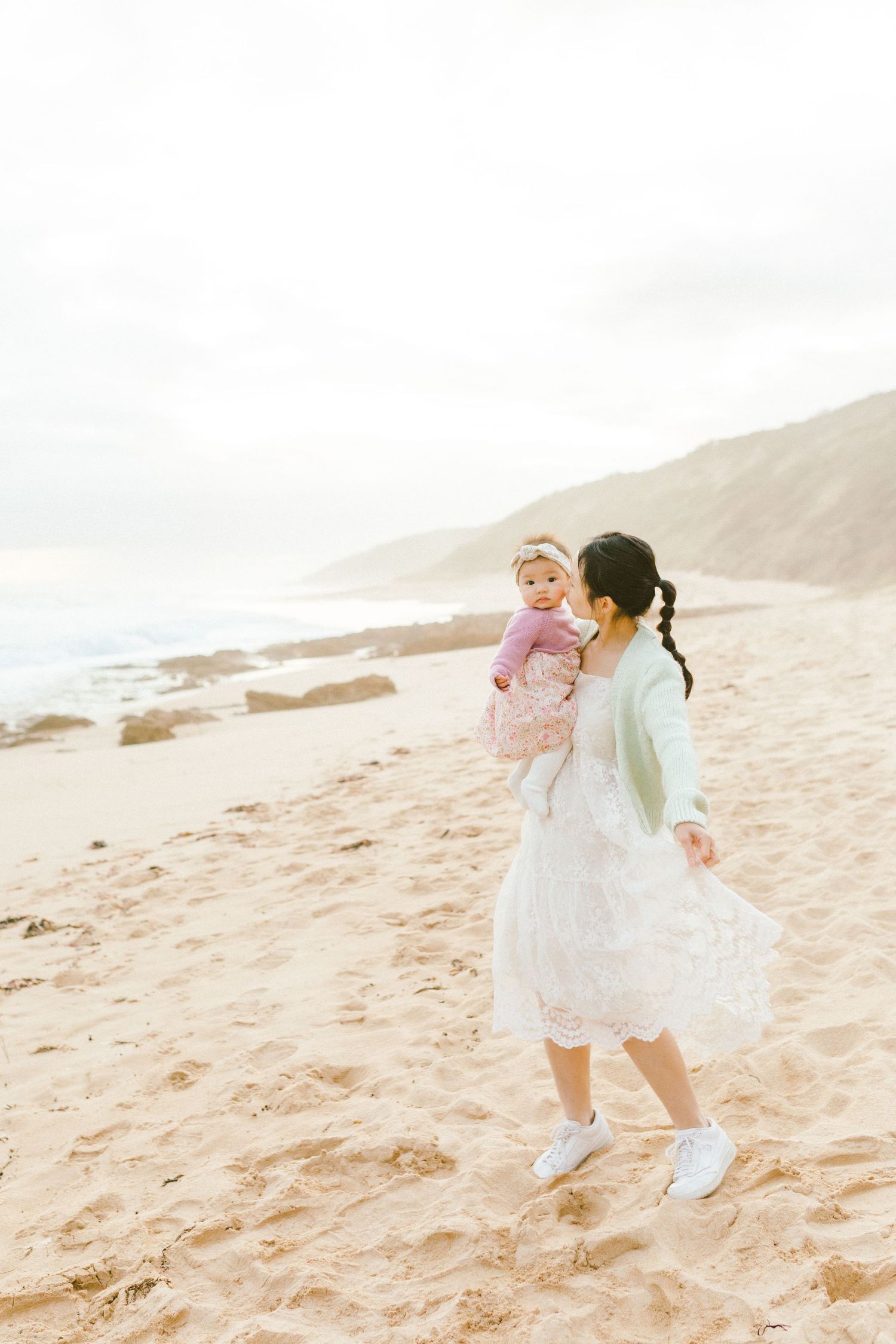 Two people in light colored outfits share a playful moment while walking along a sandy beach at sunset.
