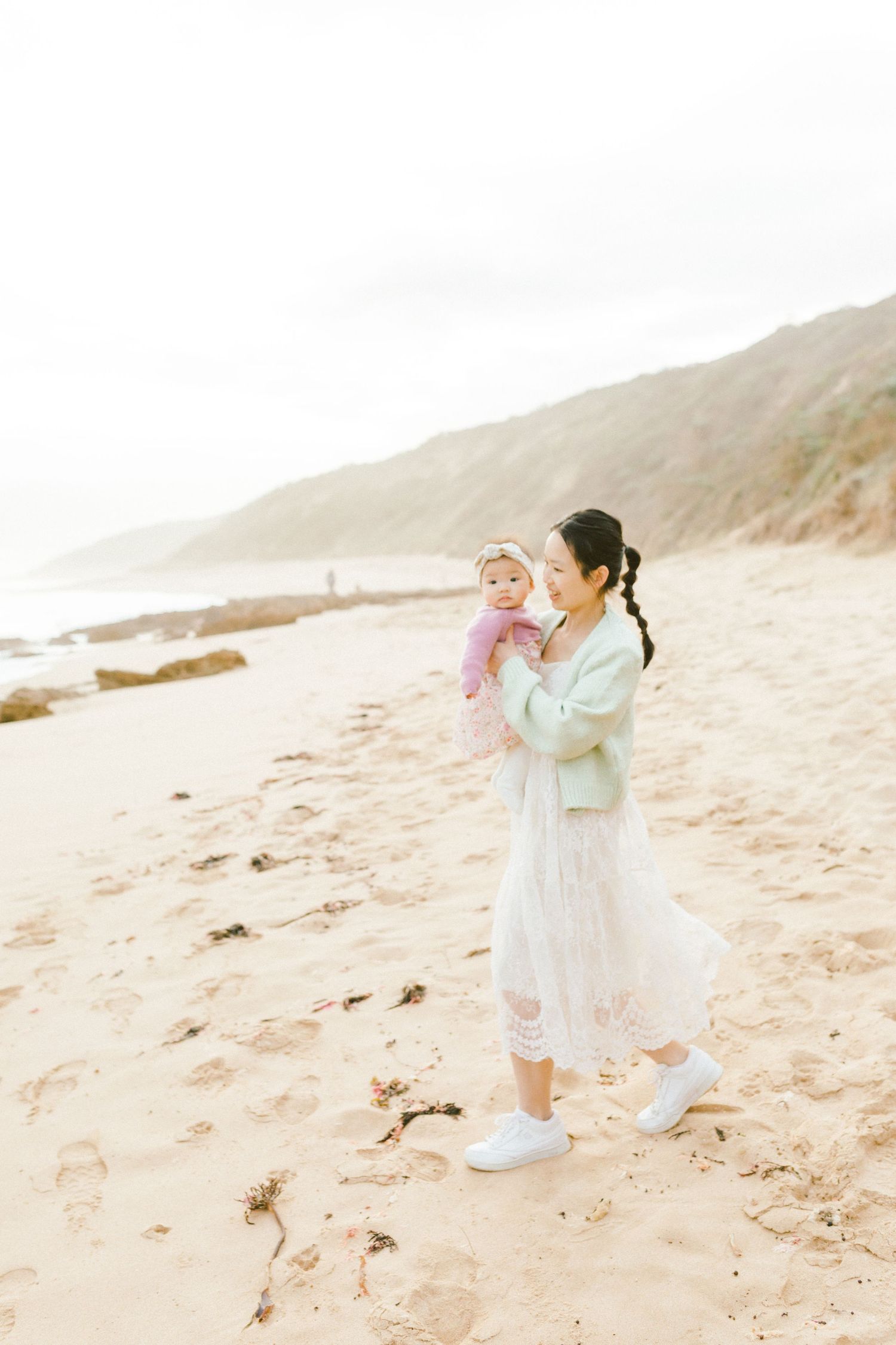 Two people in light colored clothing embrace on a sandy beach during a romantic photoshoot at sunset.