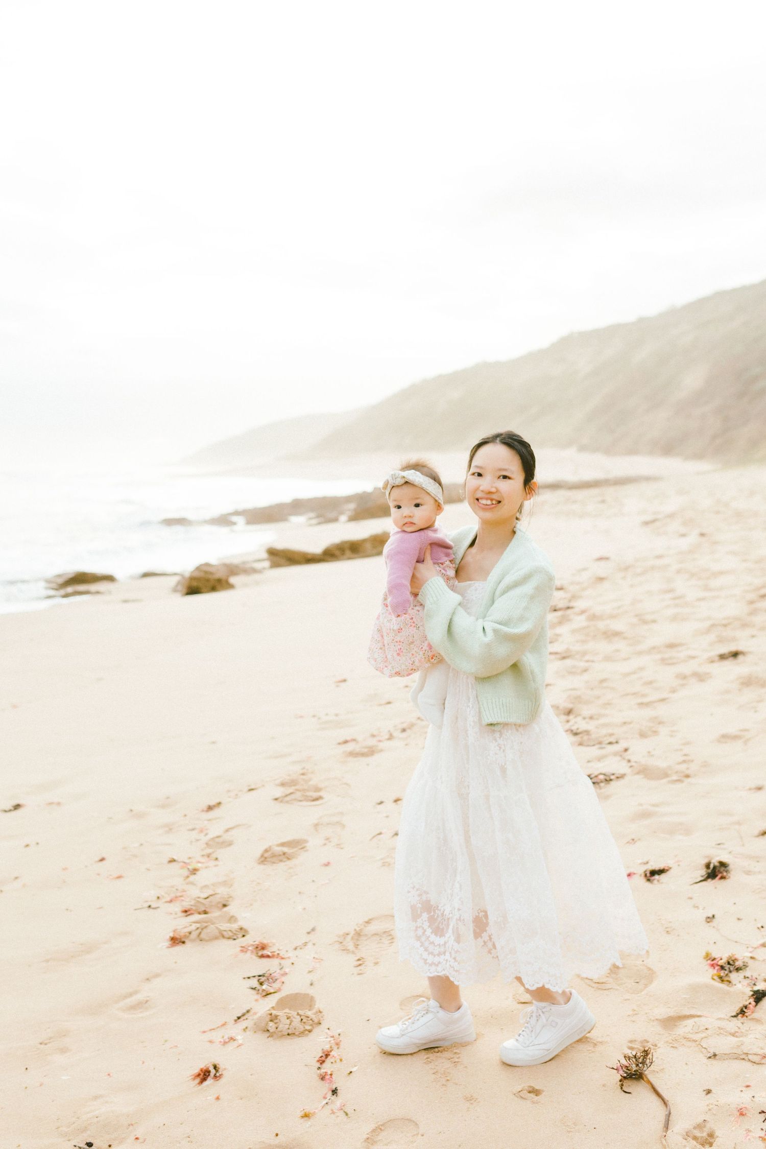 A child in a white dress plays joyfully on a sandy beach with soft, dreamy lighting and coastal scenery in the background.