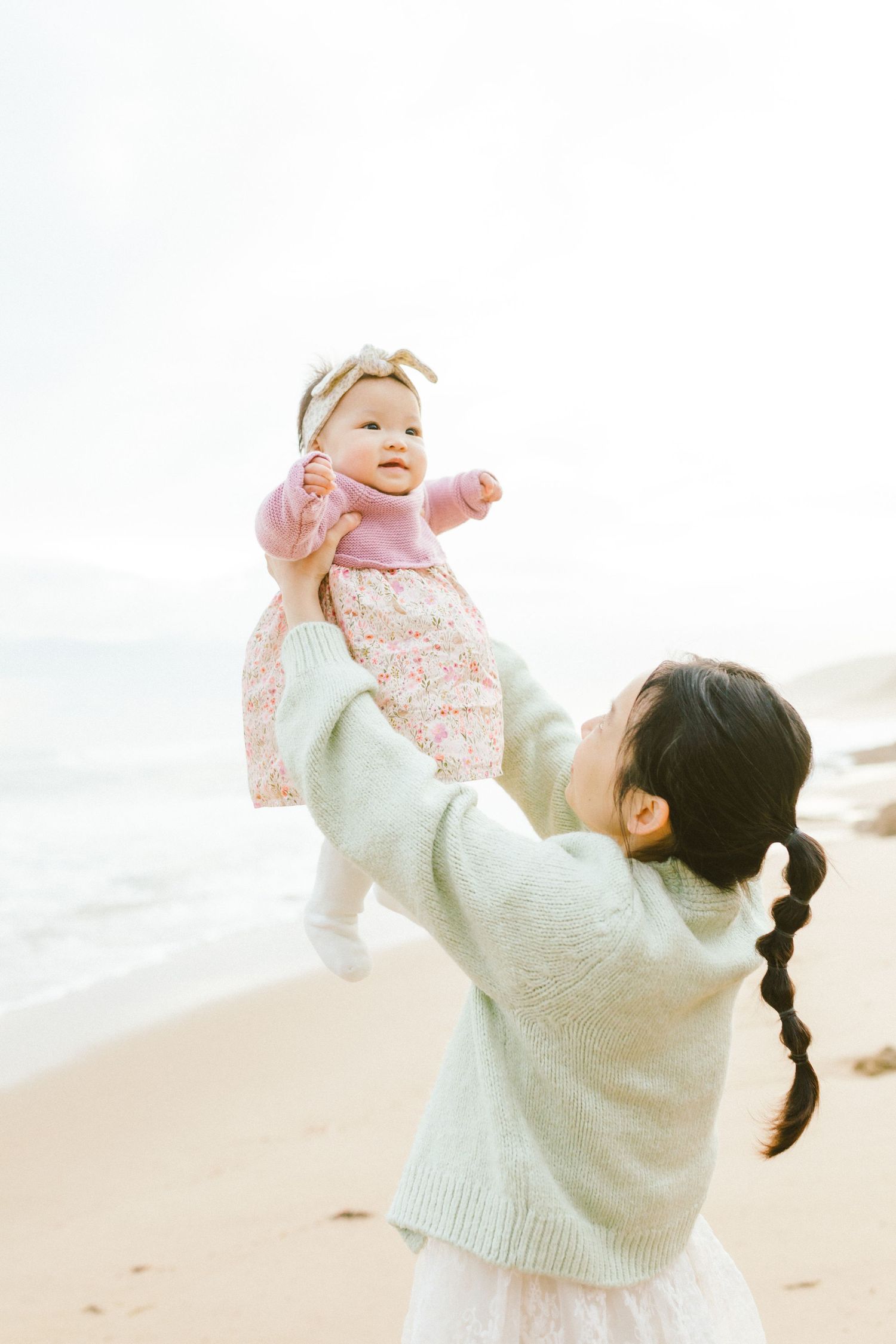 Parent in cream sweater lifts and plays with baby wearing pink on a sunny beach.