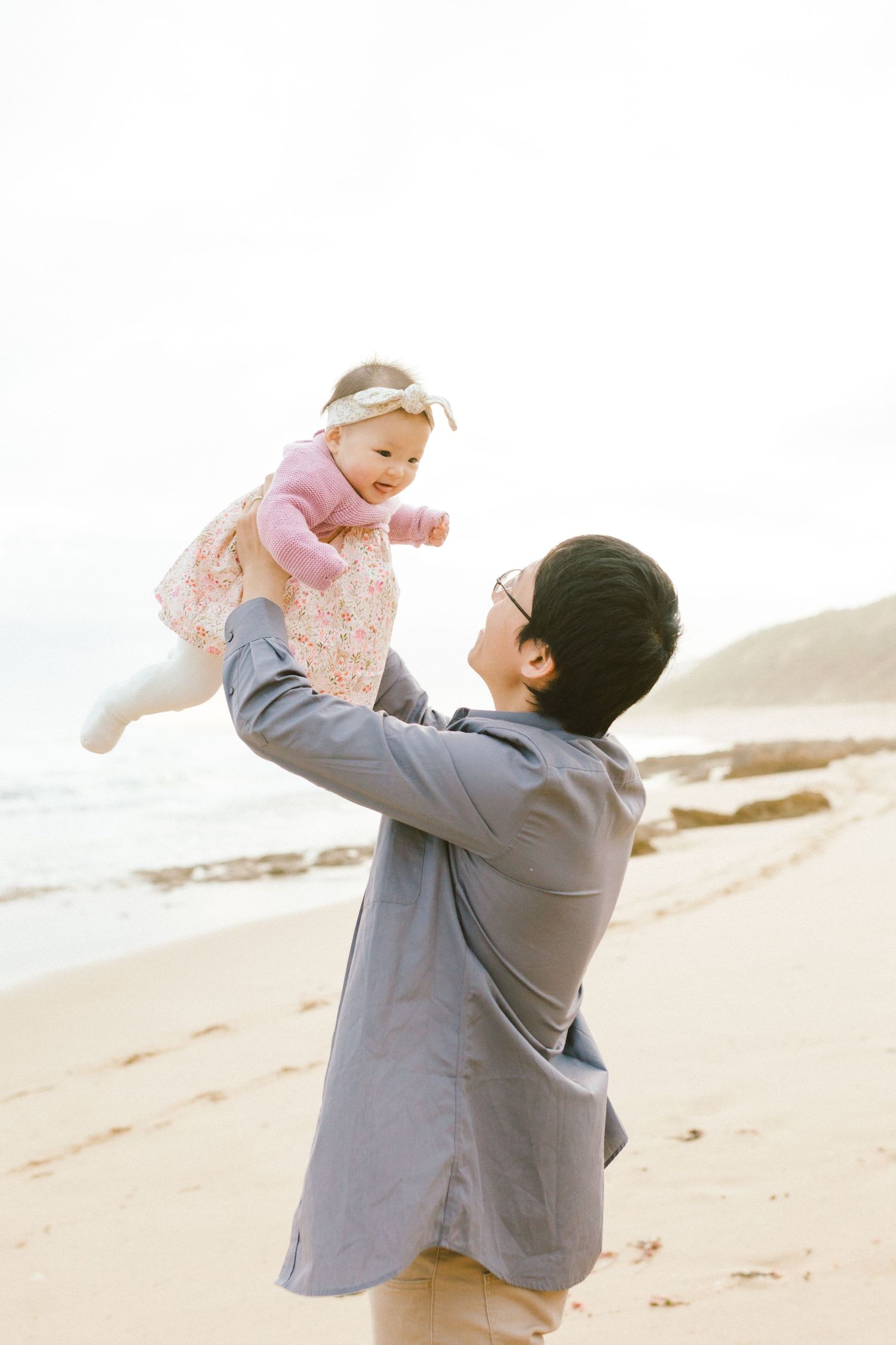 Parent in grey coat joyfully lifts baby wearing pink outfit at the beach during sunset.