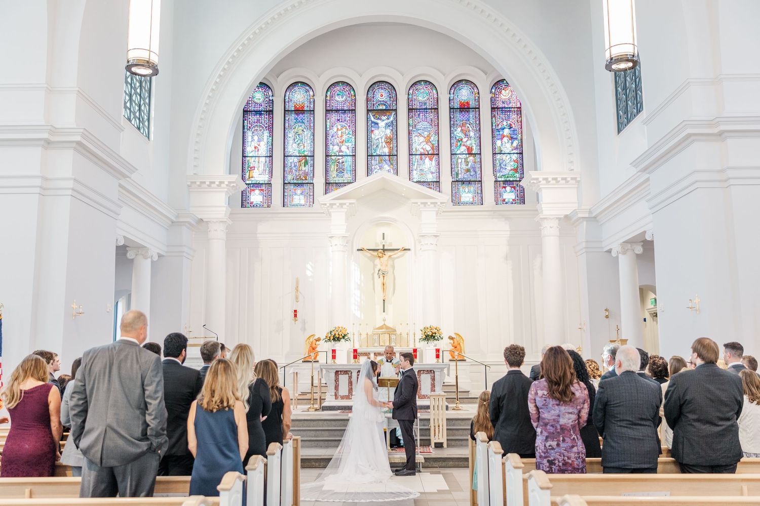 Wedding ceremony taking place in a bright white Catholic church with stained glass windows and crucifix at the altar.
