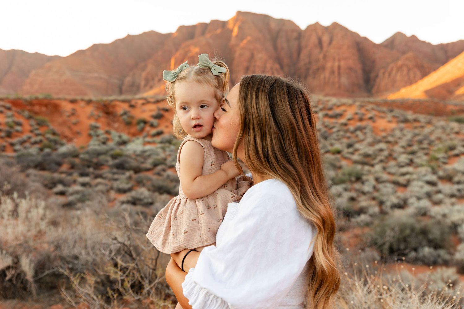 St. George Utah family photographer capturing mom kissing her 2-year-old on the cheek with red rock mountains at golden hour