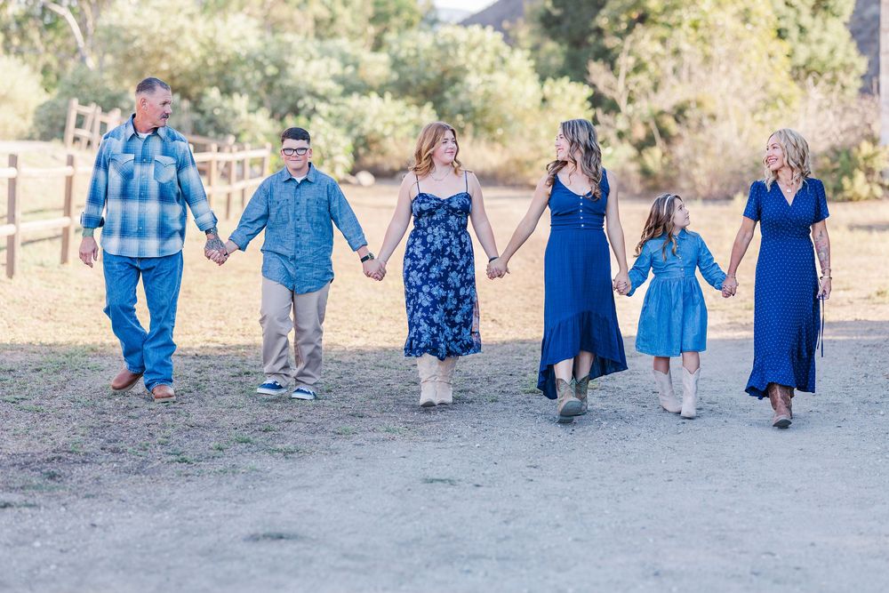 A group in coordinated blue outfits holds hands while walking on a dirt path with mountains in the background.