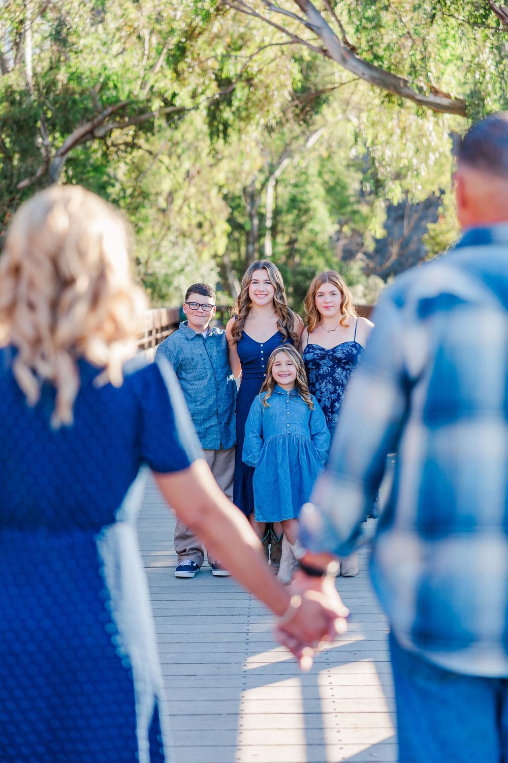 A group in coordinating blue outfits stands on a wooden boardwalk surrounded by trees on a sunny day.
