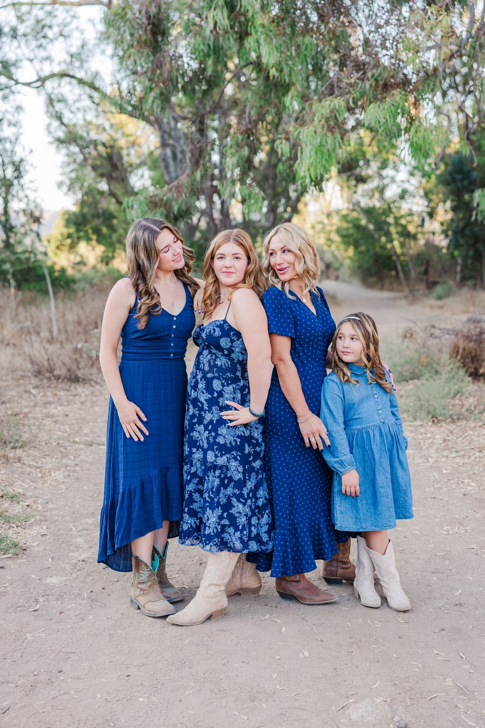 Four people wearing coordinating blue dresses sit together on a dirt path surrounded by trees.
