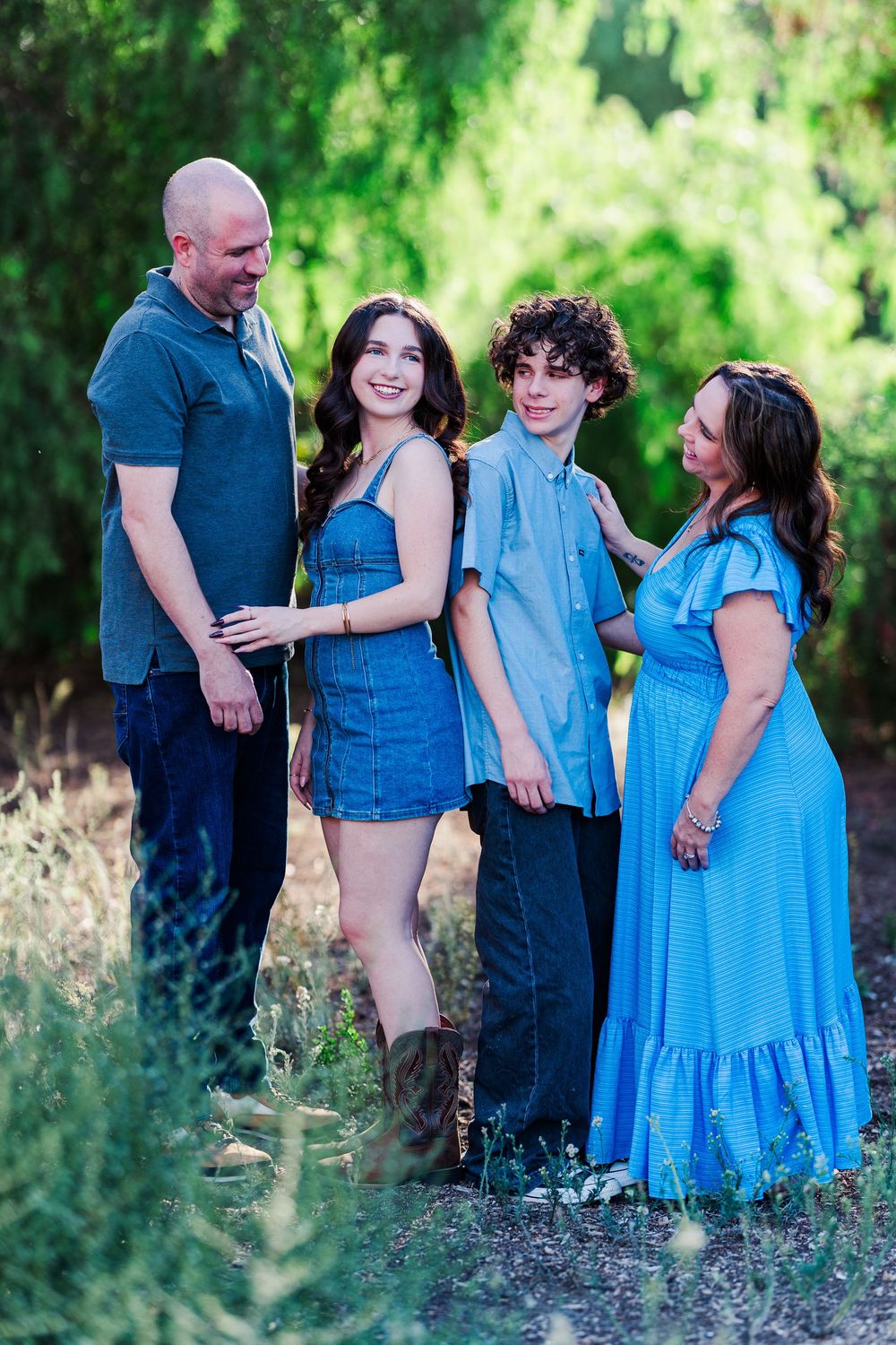 A family wearing coordinating blue outfits poses together in a natural outdoor setting with green foliage.