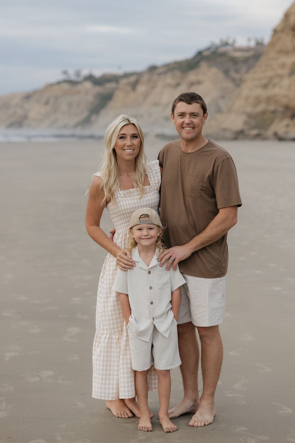 Family of three poses together on sandy beach with coastal cliffs in background during sunset photoshoot.