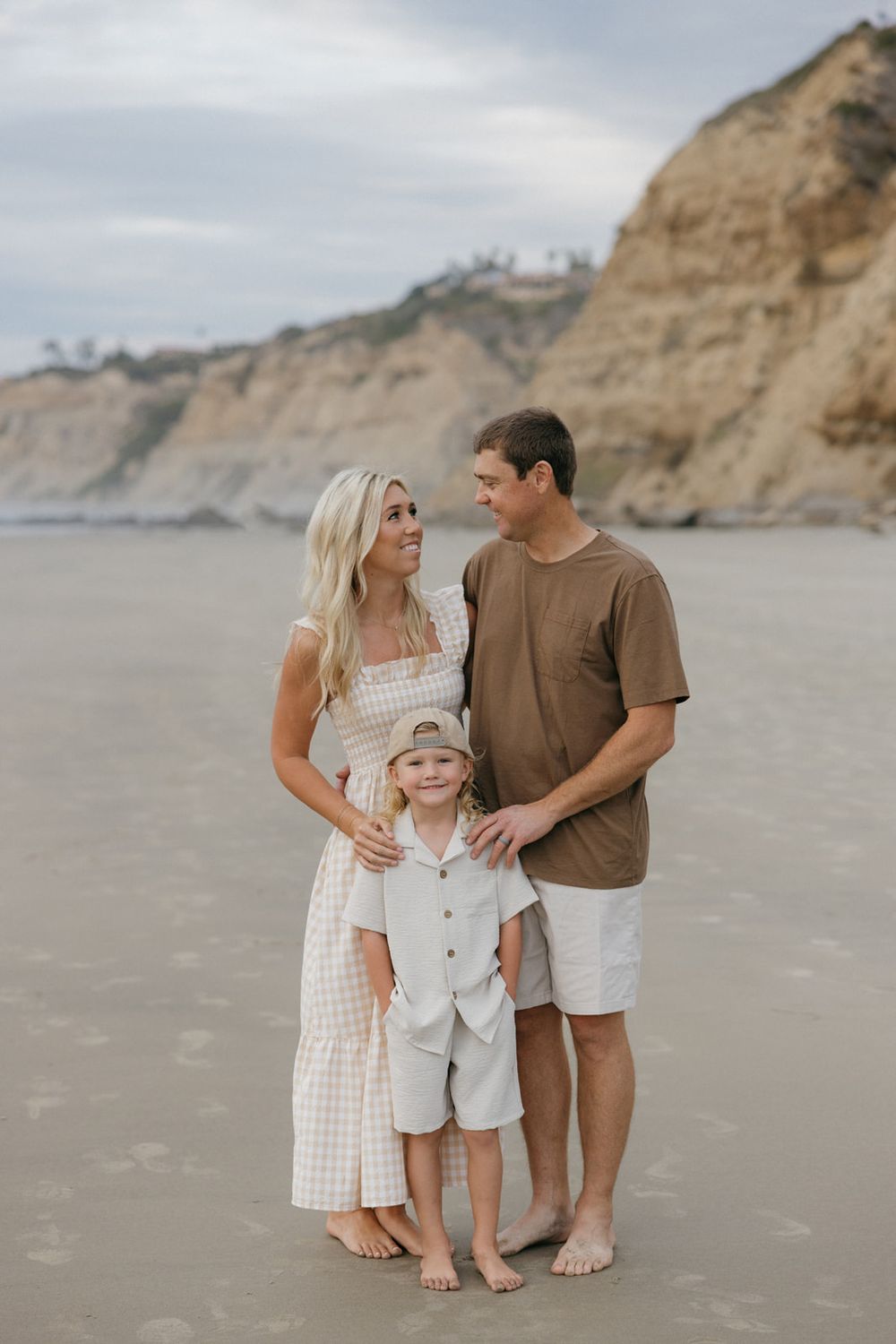 A loving family moment captured on a sandy beach with coastal cliffs in the distance.