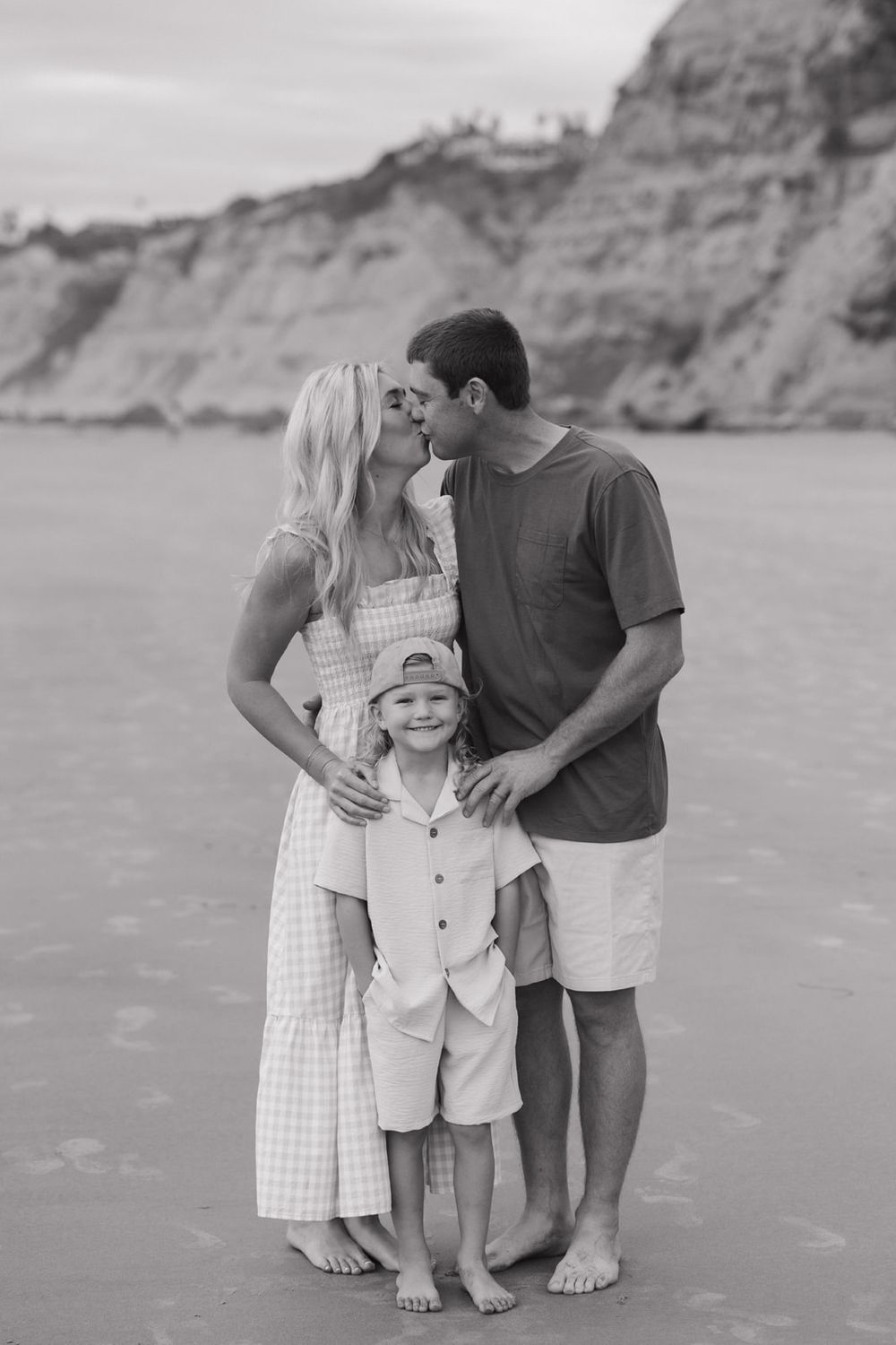 Black and white portrait of a family embracing on a beach with dramatic cliffs behind them.