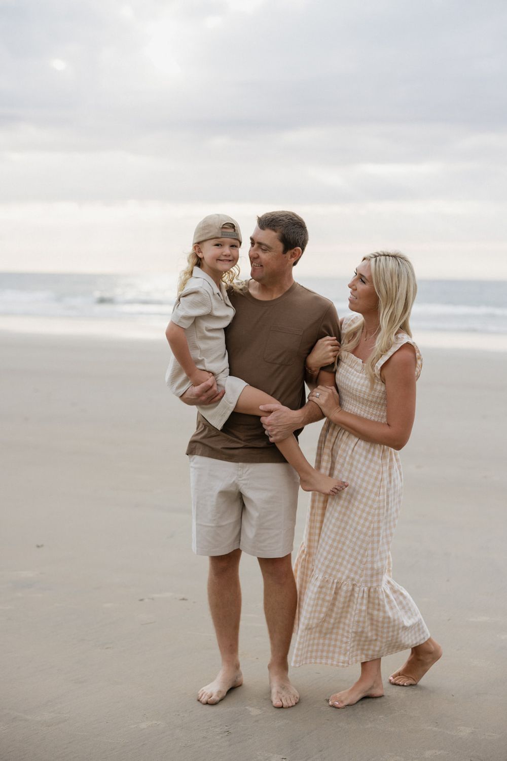 A family in neutral colored clothing shares tender moments while walking along a misty beach.