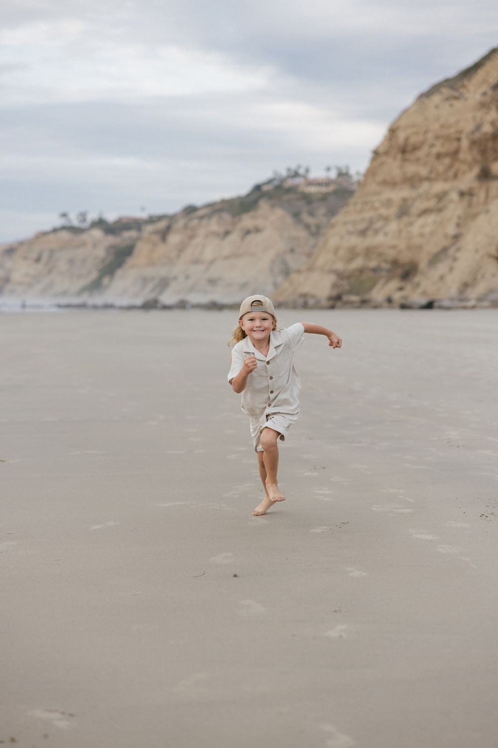 Young child in white clothing running playfully along a beach with coastal cliffs behind.