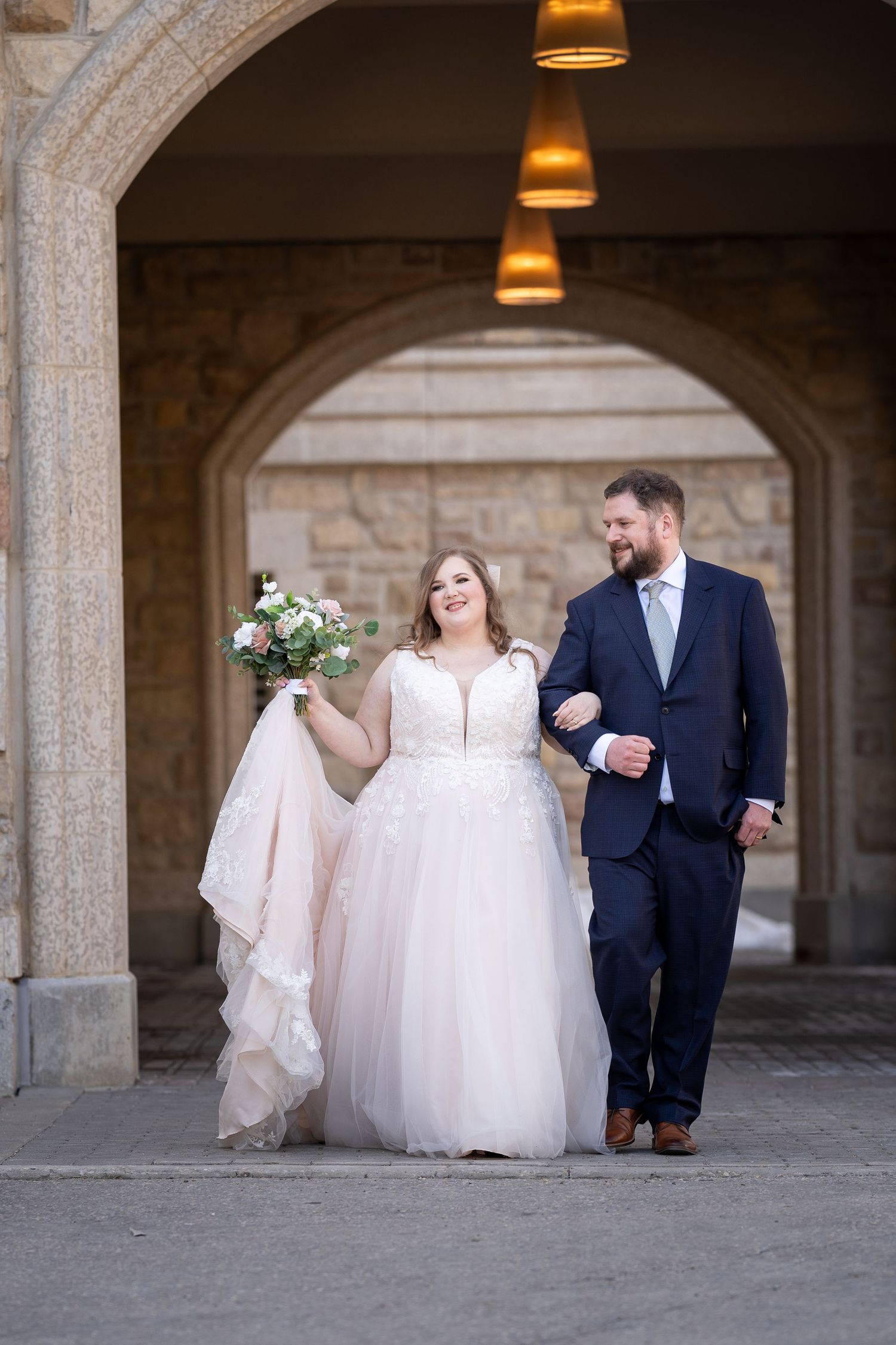 A couple walks arm in arm under stone archways, the bride in a flowing white gown holding a bouquet.