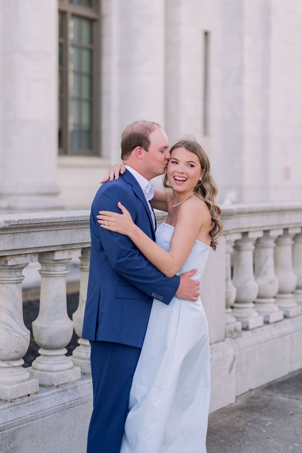 A couple embracing and laughing together against white stone architecture.