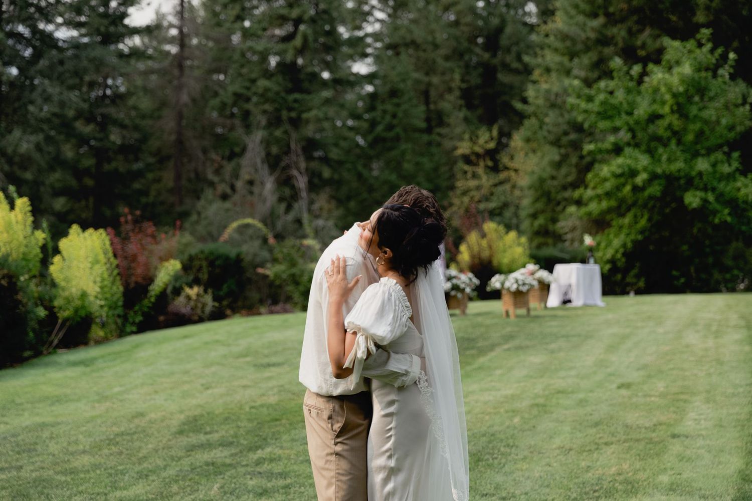 Bride and groom share a tender embrace on green lawn surrounded by lush garden landscaping.