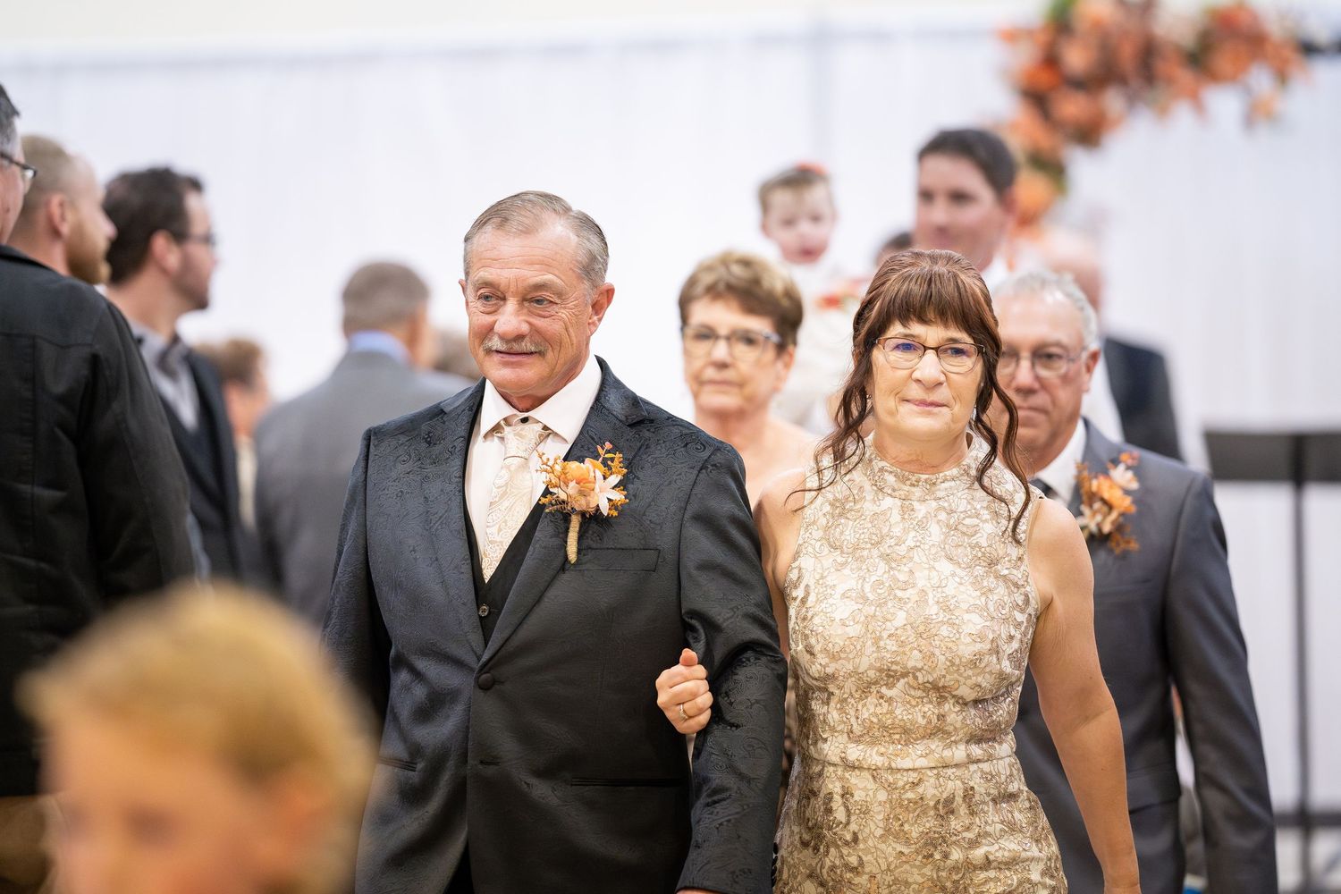 A wedding party walks down a path outdoors with guests visible in the background during an autumn celebration.