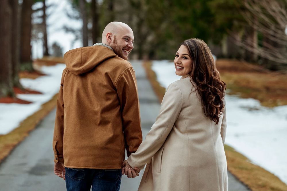 Happy couple holding hands walking on snowy path turning to smile at camera.