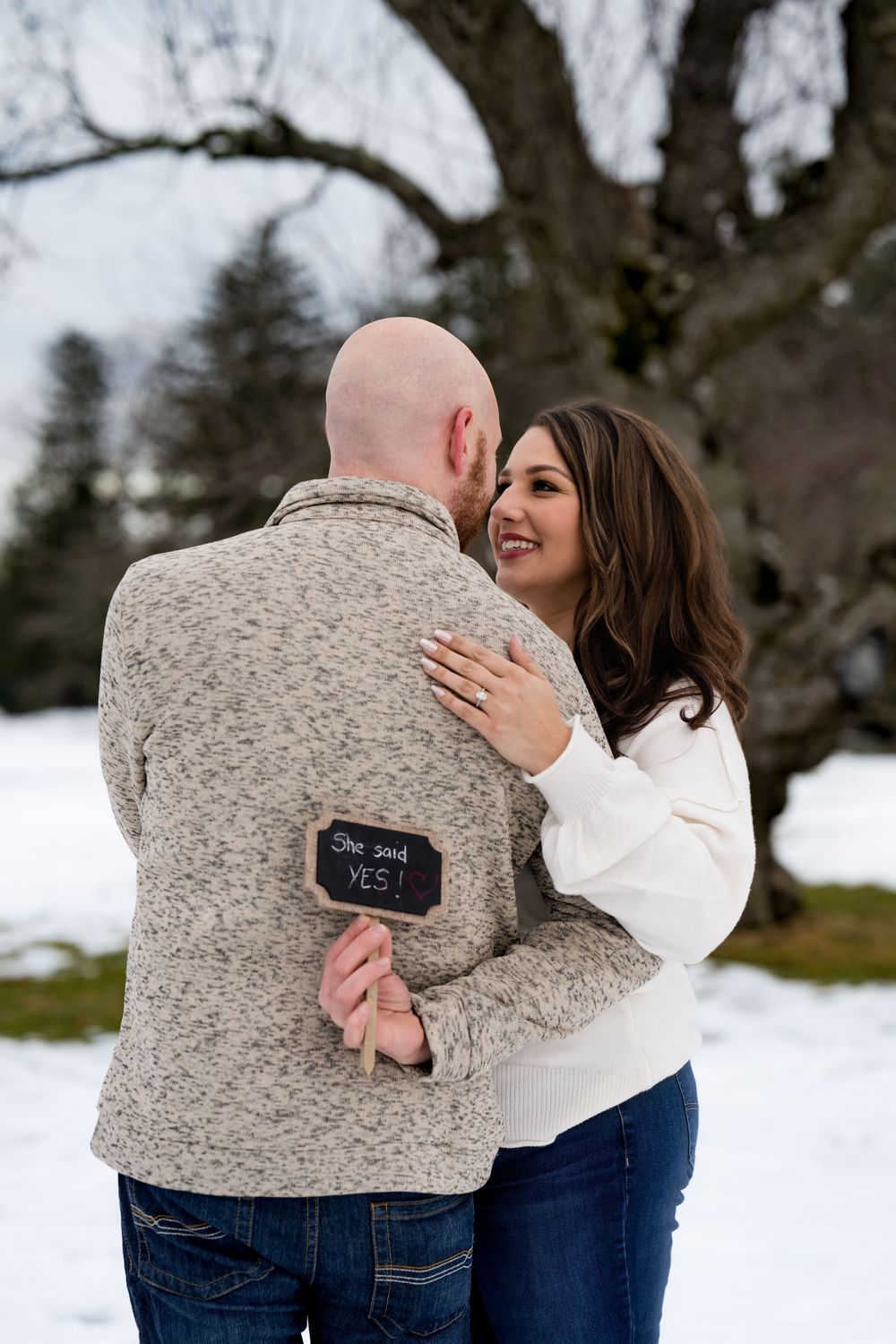 Engaged couple embracing outdoors in snow, man holding a sign reading She Said Yes.