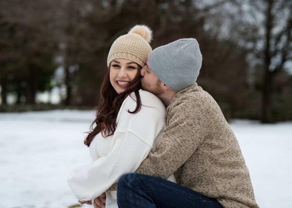 Couple sharing a tender moment outdoors in winter, woman smiling while wearing a beige knit hat.