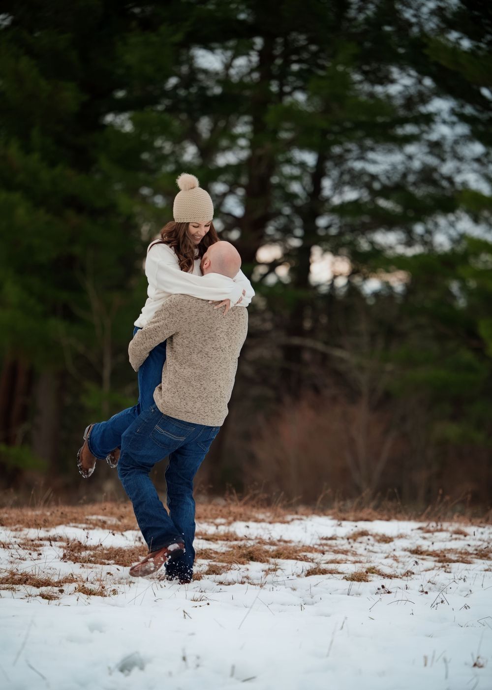 Man lifting woman joyfully in a snowy field surrounded by evergreen trees in winter.