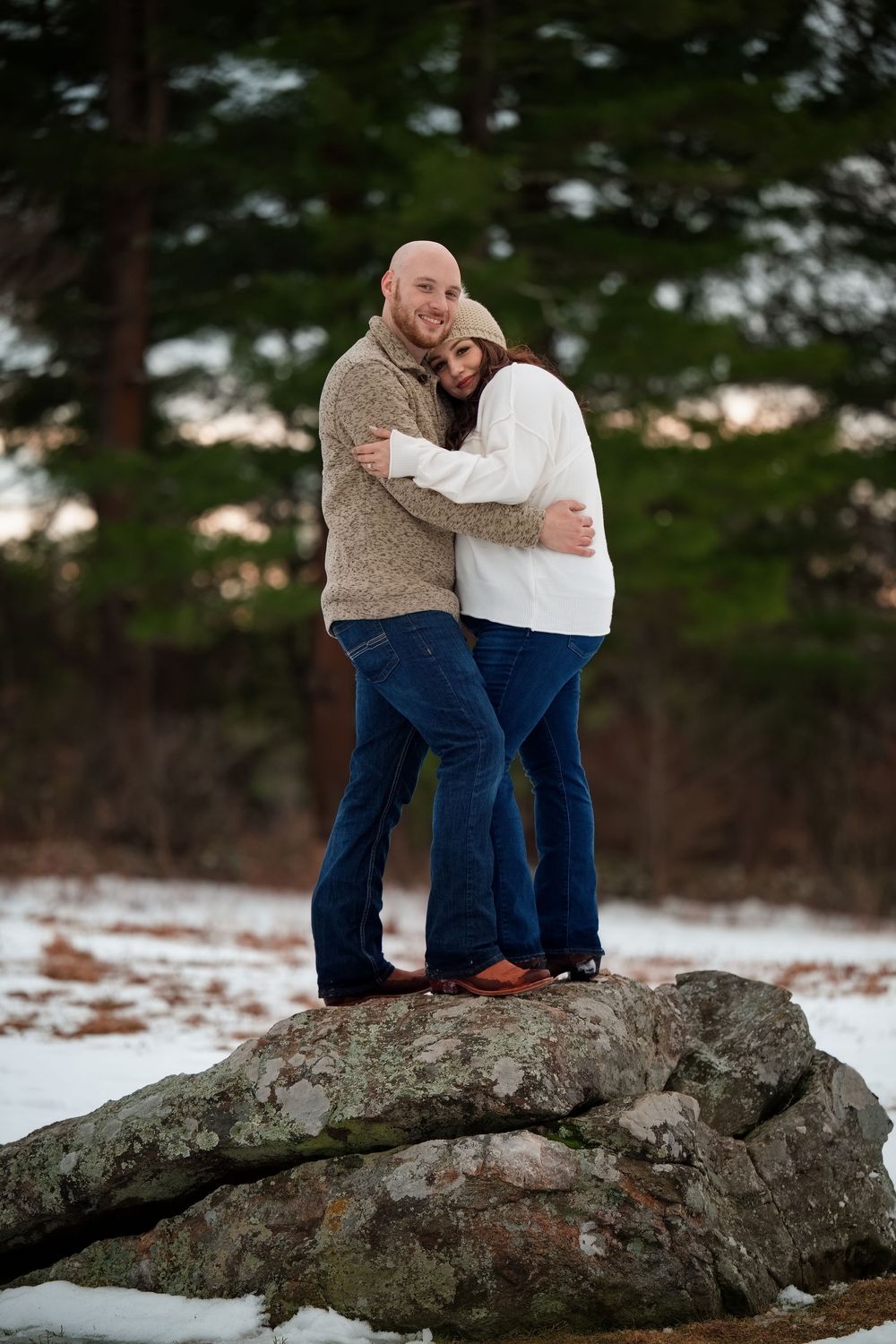 Smiling engaged couple standing together on a rock in a snowy winter forest setting.