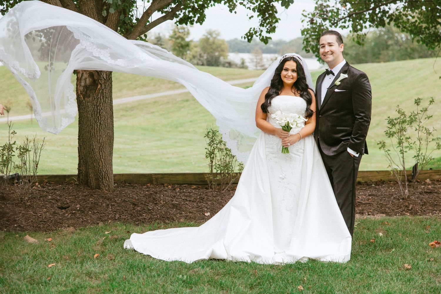 A couple in wedding attire poses outdoors on a grassy hill with the bride's veil flowing in the breeze. Metro Detroit Wedding Content