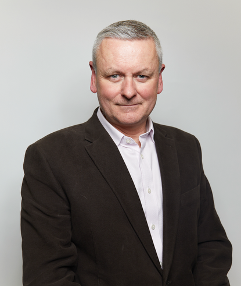 Professional headshot portrait of a person in a dark suit and white shirt against a light gray background.