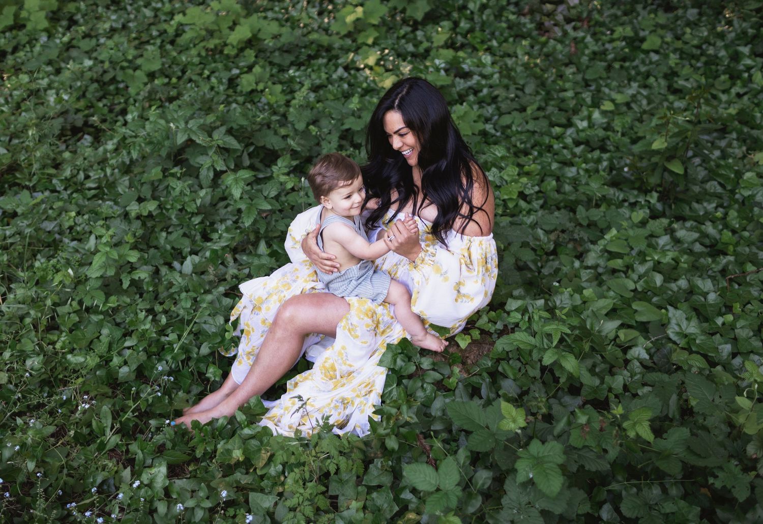 A happy mother with long dark hair in a floral gown holds her baby boy on her lap in lush greenery for a motherhood session. MA motherhood