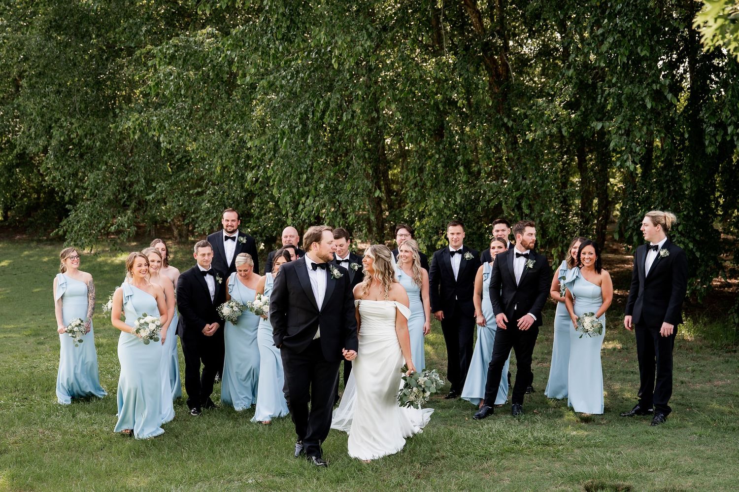 Wedding party in light blue dresses and black tuxedos poses together on a lush green lawn.