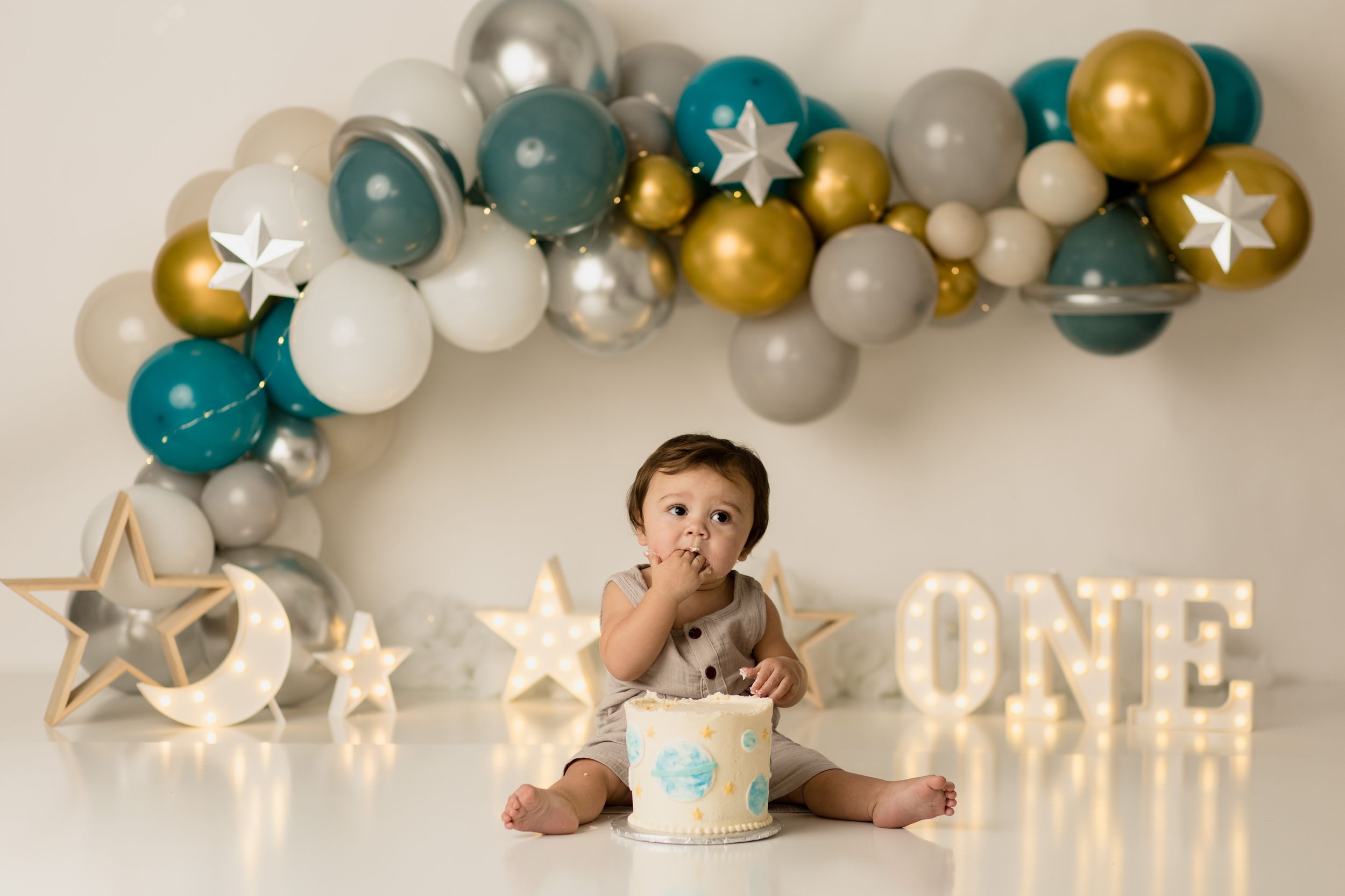 Balloon arch in teal, gold, and white colors with illuminated star decorations and a 'ONE' light sign display.