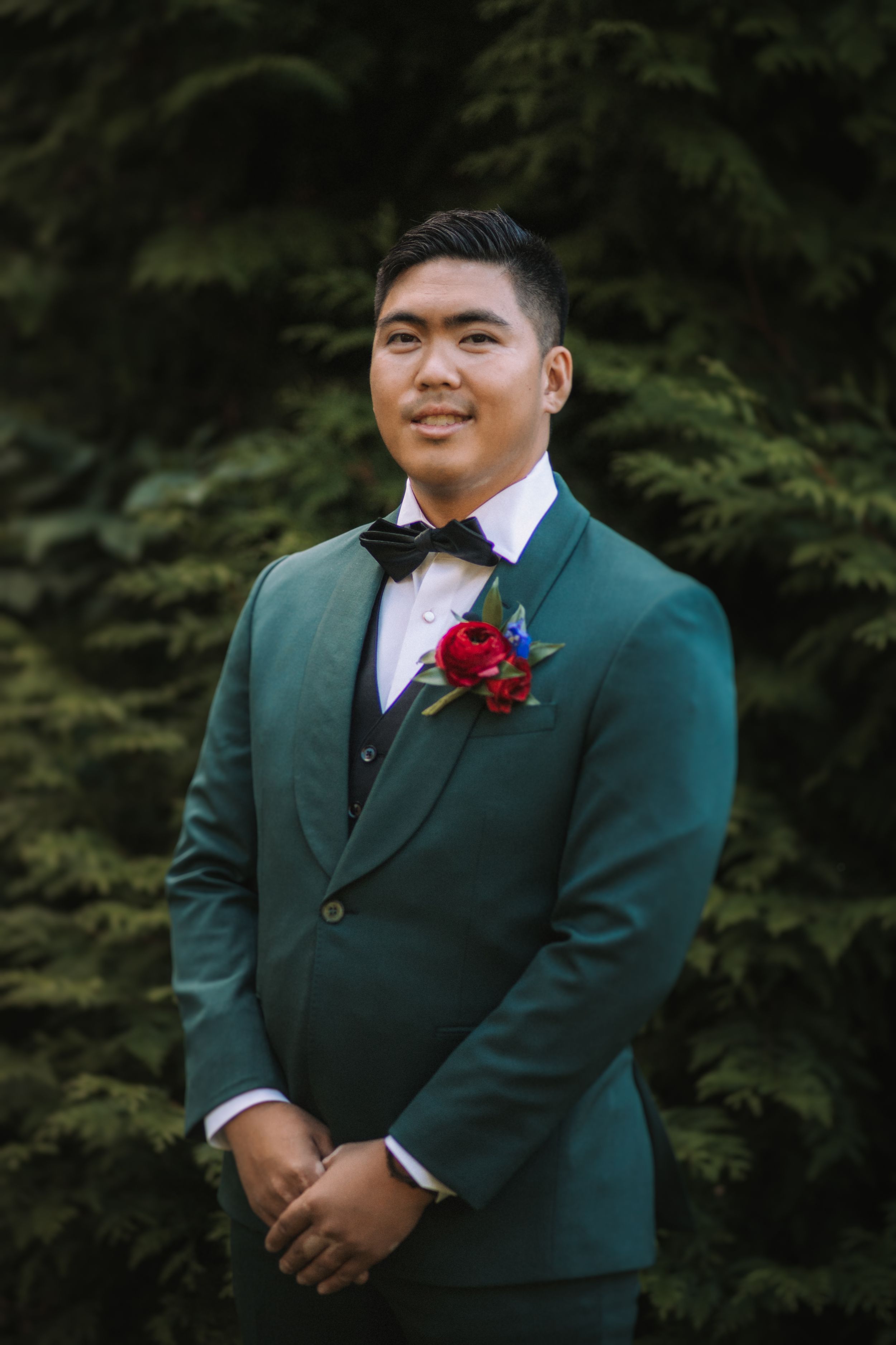 Wedding party member wearing a dark green suit jacket with a red boutonniere poses outdoors against greenery.